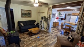 A cozy living room with a dark sofa, wooden coffee table, and a fireplace. A dining area with a table and chairs is visible through an open doorway. - Sleeping Wolf Guest House (Oakhurst)