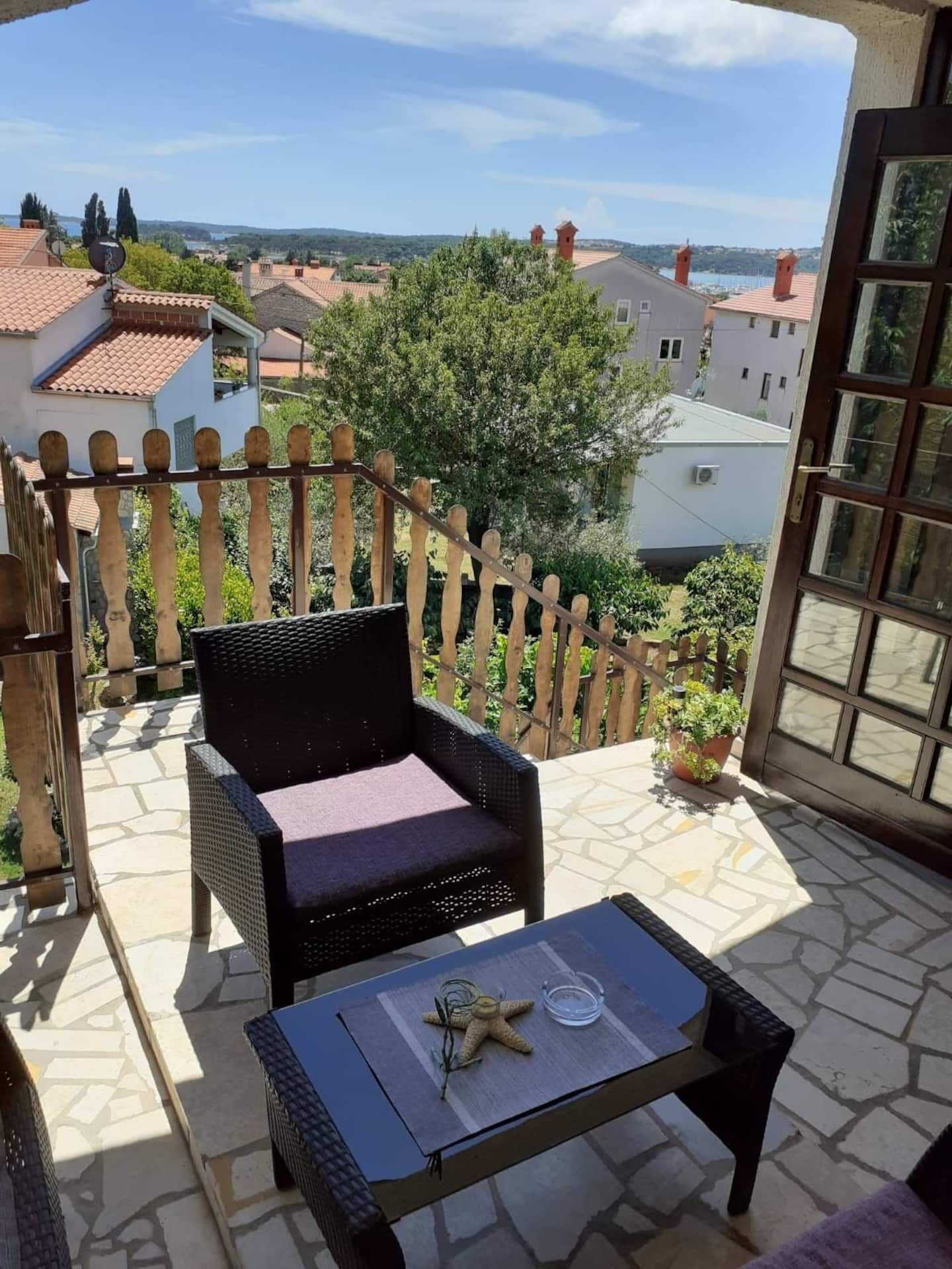 A balcony with a wicker chair and a small table, overlooking a residential area with houses and trees.