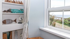 Une pièce avec un mur blanc, un banc en bois et une fenêtre donnant sur un jardin. On peut également apercevoir des étagères ornées d’objets décoratifs et un rideau.