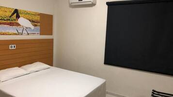 A hotel room with a bed, an air conditioning unit, a window with a blind, and a wall-mounted picture of a bird.