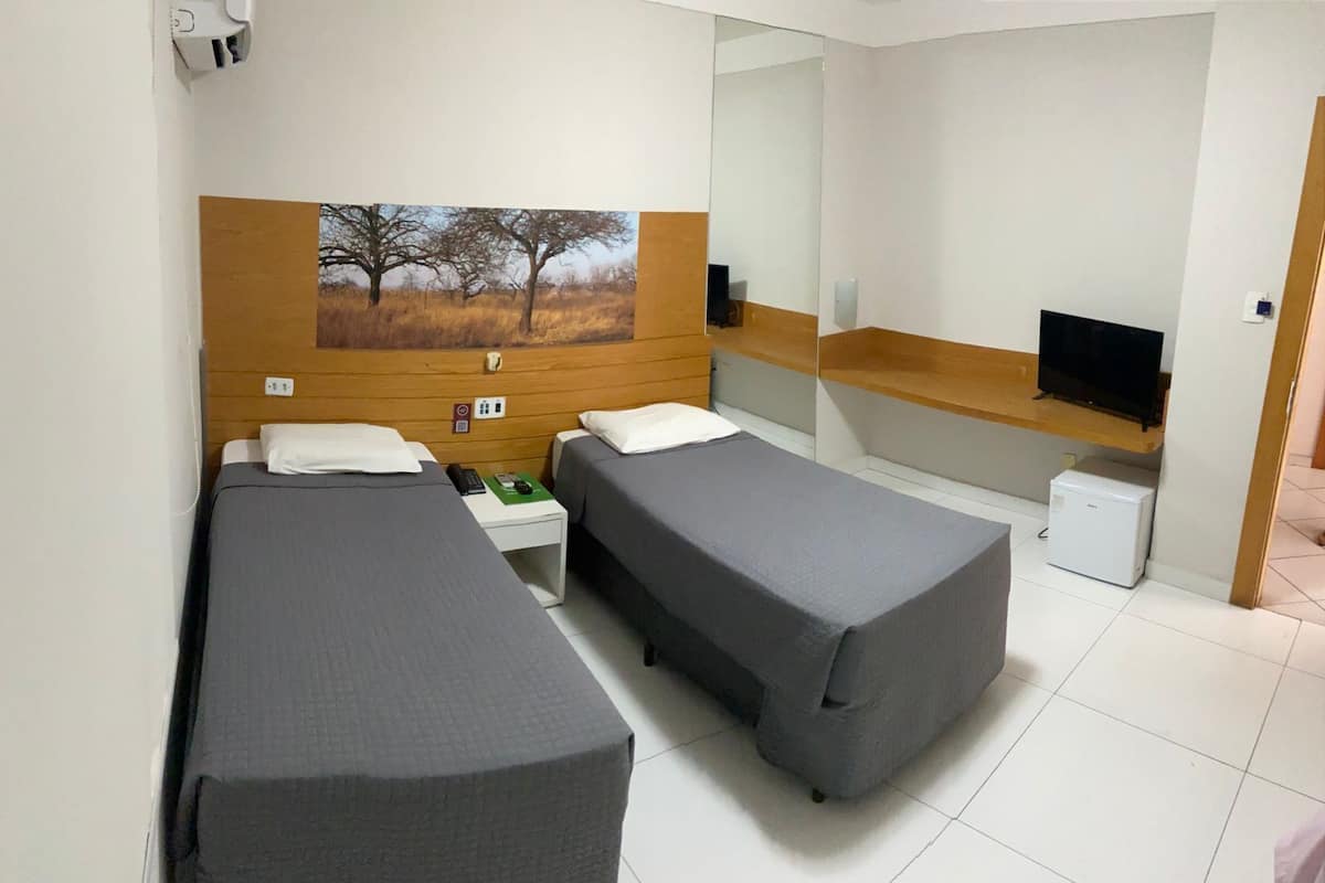 A hotel room with two single beds, a wooden headboard, a flat-screen TV, a small refrigerator, and a painting of trees.