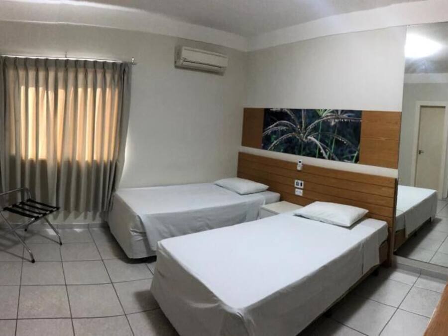 A hotel room with two single beds, a wooden headboard, a small table, a chair, a window with curtains, and an air conditioning unit.