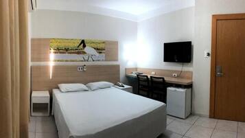 Quarto de hotel com cama, escrivaninha, cadeira, televisão e porta.
