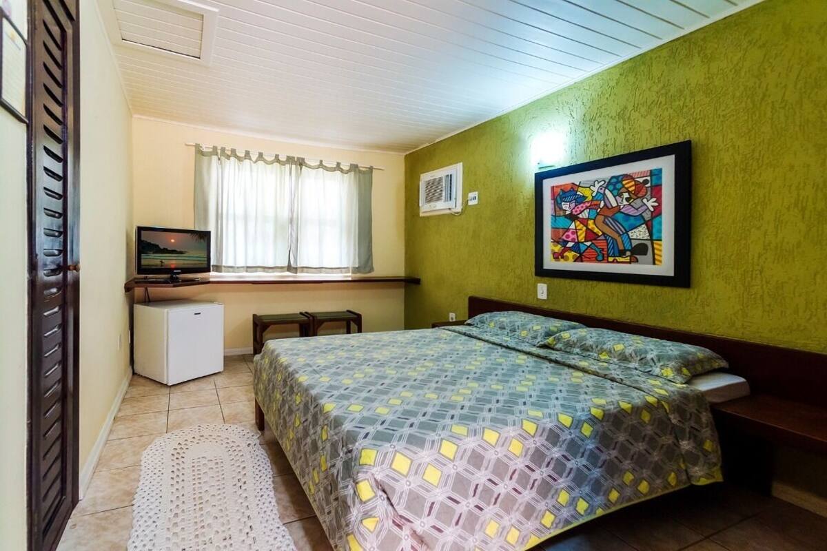 Ein Schlafzimmer mit Bett, Fernseher, kleinem Kühlschrank und einem Fenster mit Vorhängen.