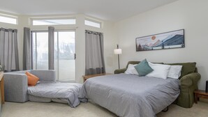 Une chambre moderne avec un grand lit, un canapé-lit et un bureau. Il y a une fenêtre avec des rideaux, un tableau au mur et une lampe sur le bureau.