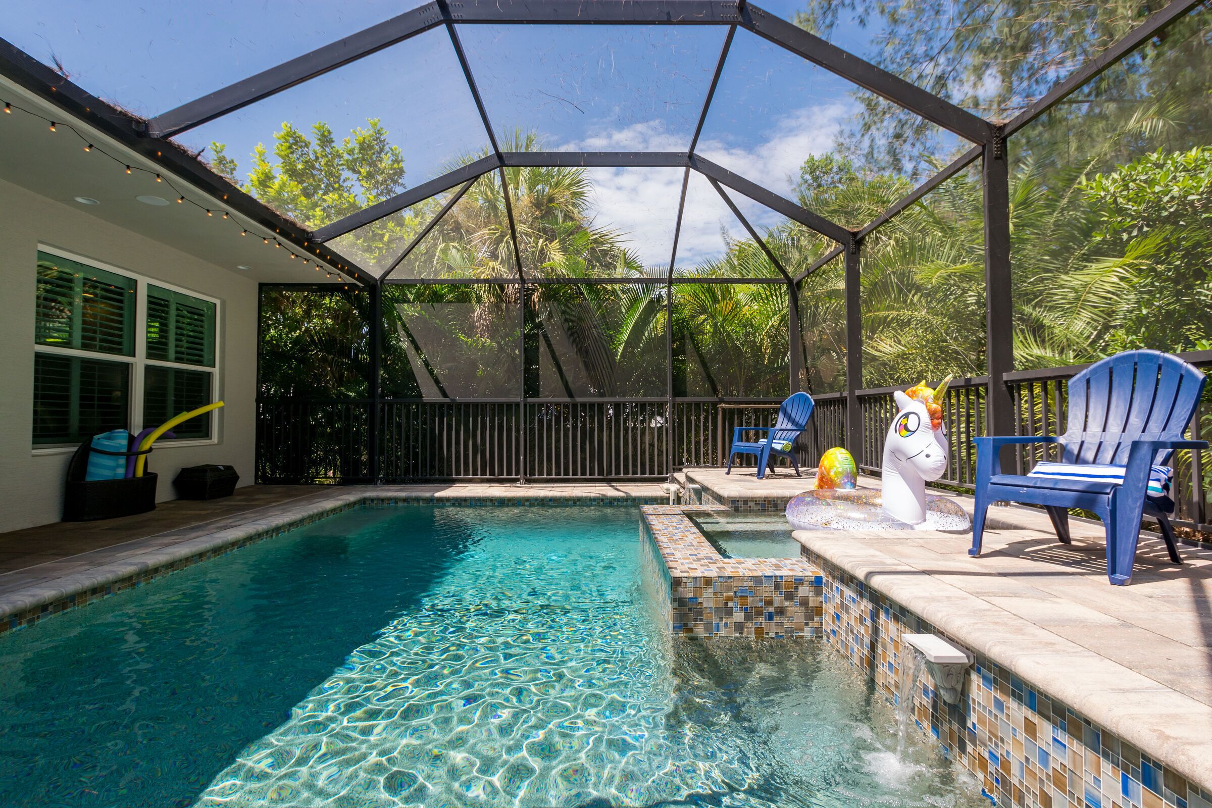 A swimming pool with a tiled edge, surrounded by a covered patio area with blue chairs and a decorative sculpture.