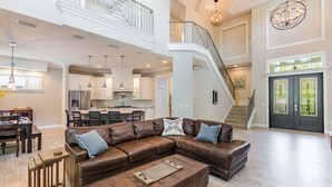 A spacious living area with a large brown leather sectional sofa, a wooden coffee table, and a dining table with chairs. A kitchen is visible in the background, and there is a staircase leading to an upper level. - The Seagrape Villa -Family Friendly- Luxury Accommodation in Paradise (Sarasota)