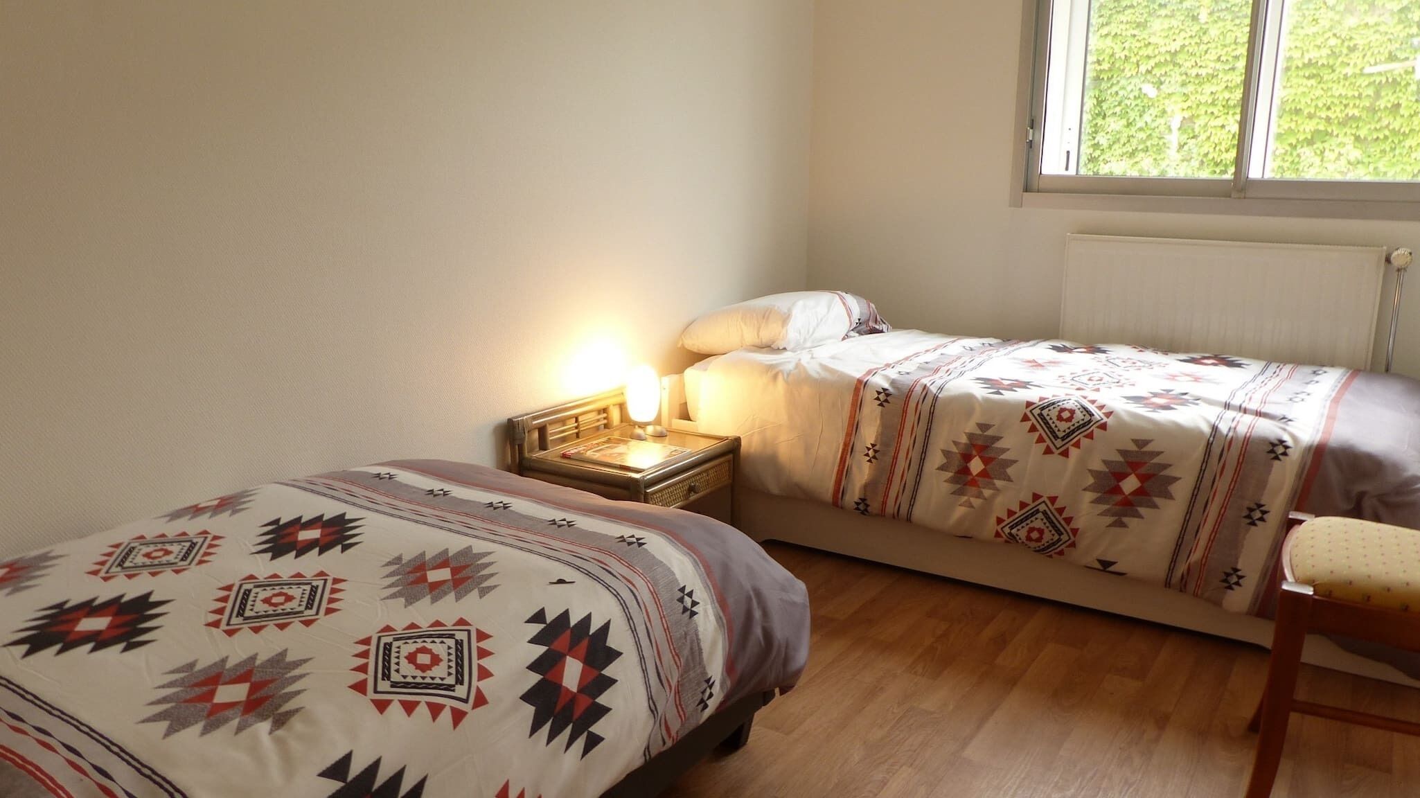 Een slaapkamer met twee eenpersoonsbedden, een nachtkastje met een lamp, een stoel en een raam met uitzicht op groen.