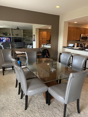 A modern dining area with a glass-top table surrounded by leather chairs, a kitchen in the background, and a TV displaying a news broadcast. - Beautiful Grayhawk Condo! Negotiated rates and FLEXIBLE Dates-contact owner (Scottsdale)