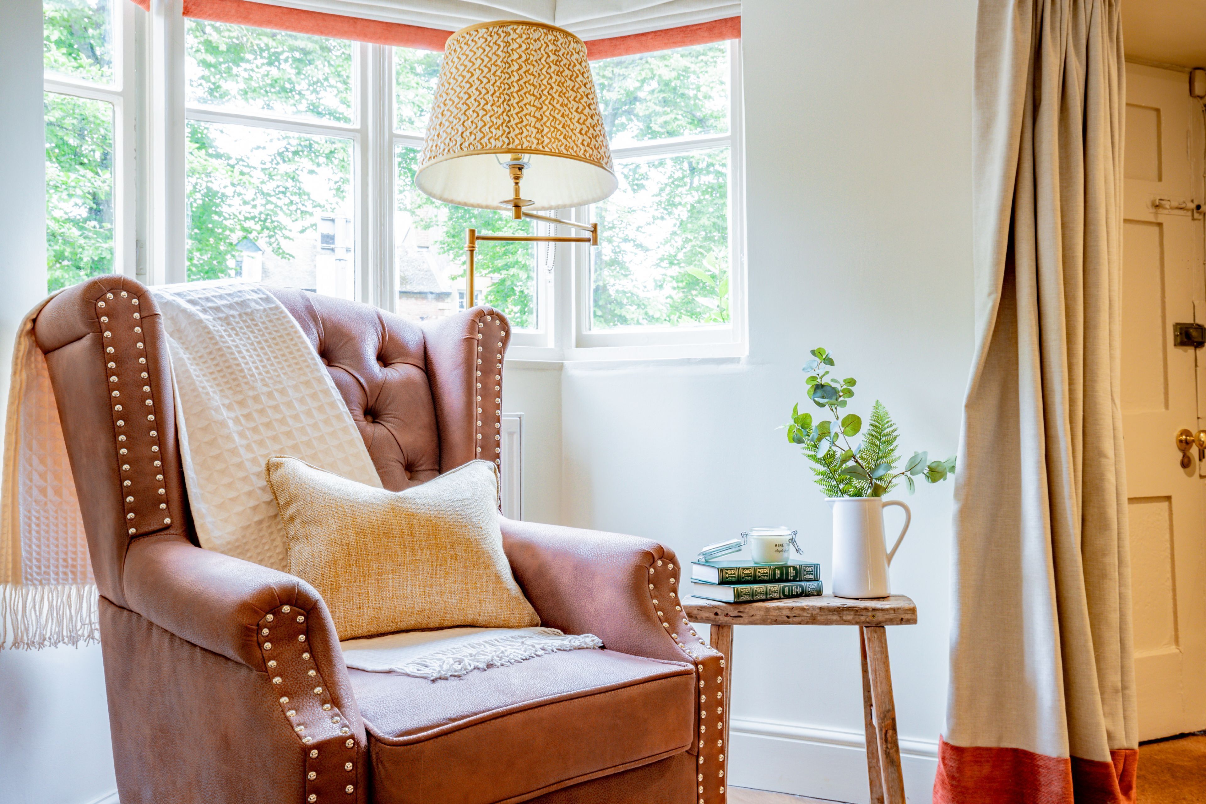 A brown leather armchair with a cushion, a small wooden side table with books and a plant, and a window with a view of greenery.
