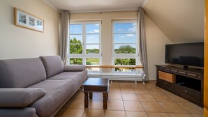Ett vardagsrum med en soffa, ett soffbord, en tv och stora fönster med utsikt över grönområden.