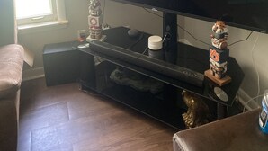 A living room with a flat-screen TV on a stand, a coffee table, a couch, and a window with blinds. - A great peaceful apartment (Mississippi Biloxi)