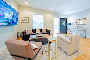 A modern living room with a dark sofa, two armchairs, a coffee table, and a flat-screen TV mounted on the wall displaying various channels.