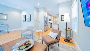 Una sala moderna con un sofá, una mesa de centro con un plato decorativo, una zona de comedor con una mesa y sillas, y una cocina al fondo.