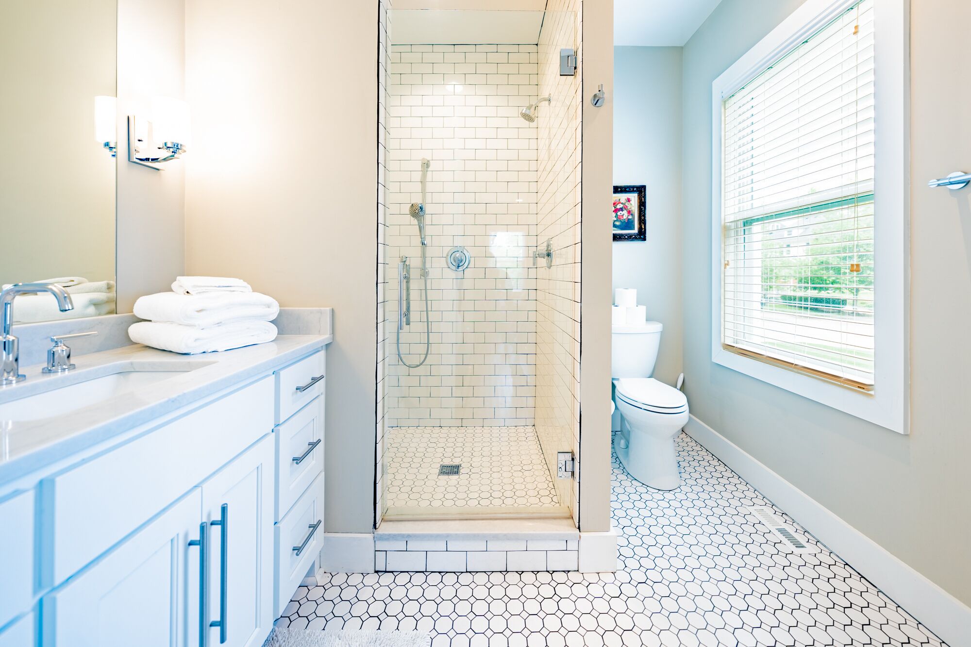 A modern bathroom with a glass-enclosed shower, a white sink with a marble countertop, and a toilet with a window view.
