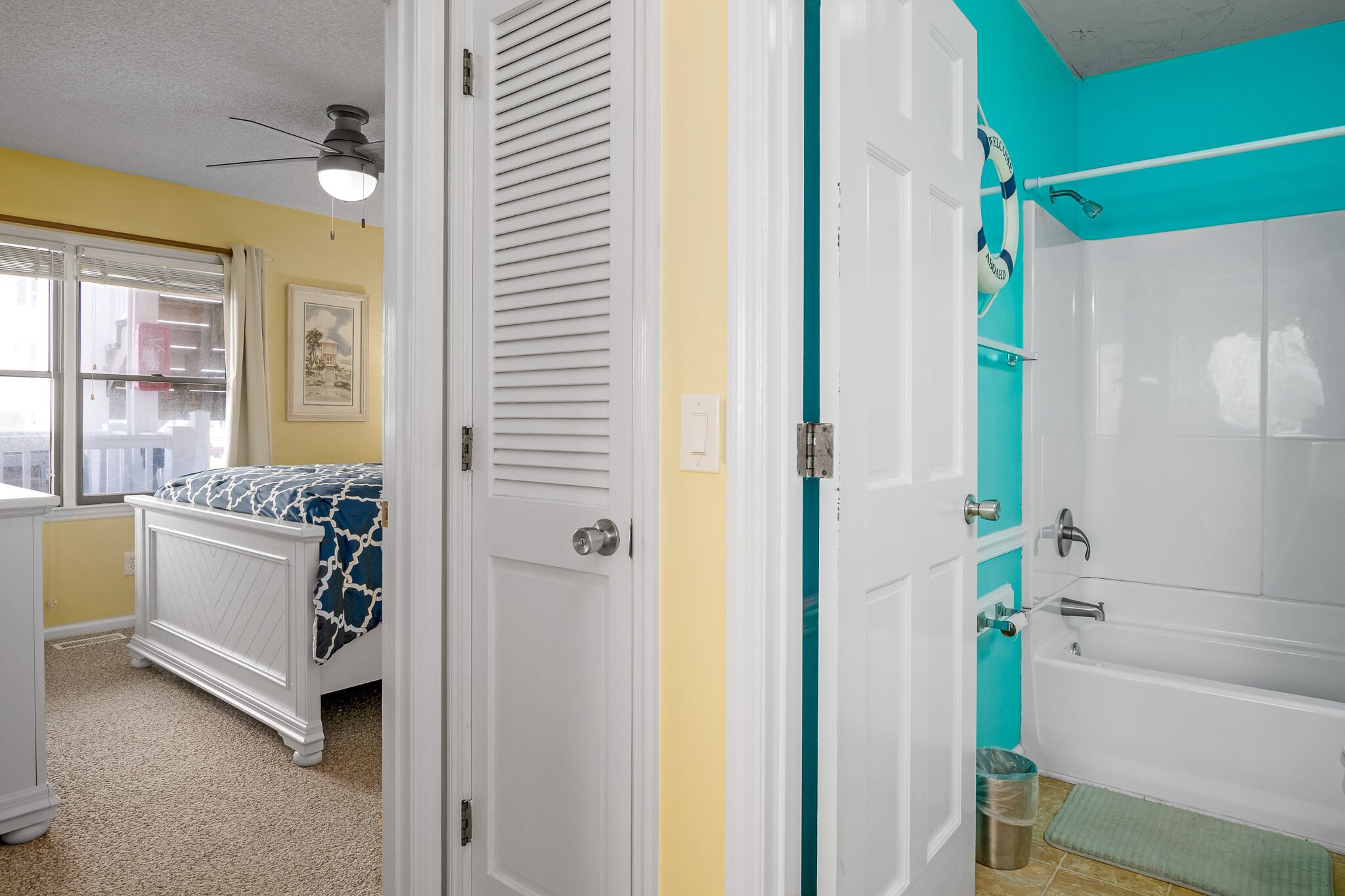 Un dormitorio con cama, ventana, ventilador de techo y puerta que da a un baño con bañera y ducha.