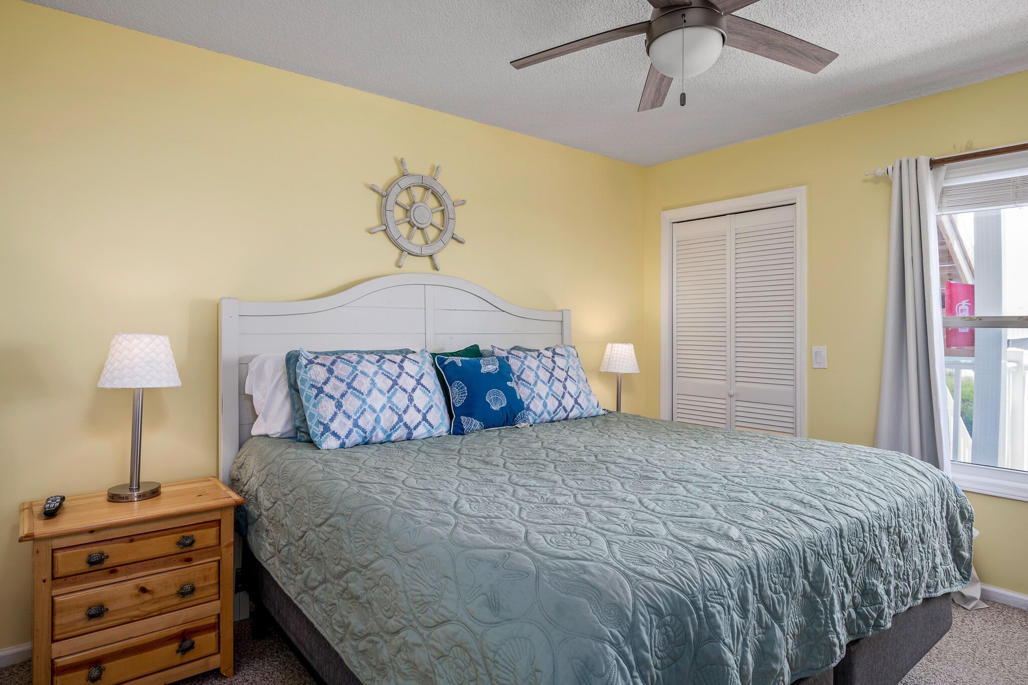 A bedroom with a bed, a nightstand, a ceiling fan, a window with curtains, and a wall clock.