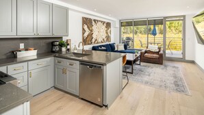 A modern kitchen with grey cabinets, a dishwasher, and a coffee maker. There is a dining area with a blue sofa and a leather chair. A large window provides a view of the outdoors.
