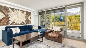 A modern living room with a blue sofa, a brown leather chair, a wooden coffee table, and a large window offering a mountain view.