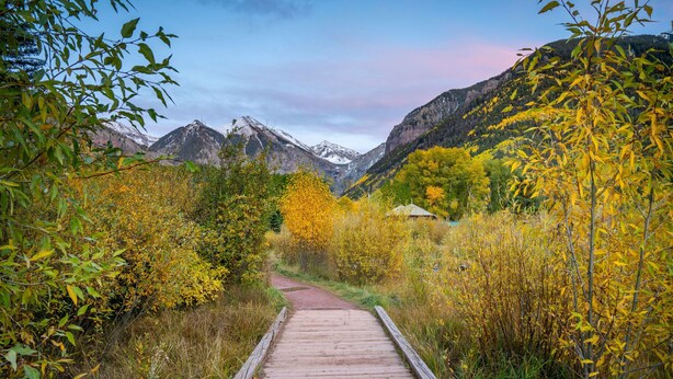 A wooden pathway leading through a mountainous area with autumn foliage and snow-capped peaks in the background.