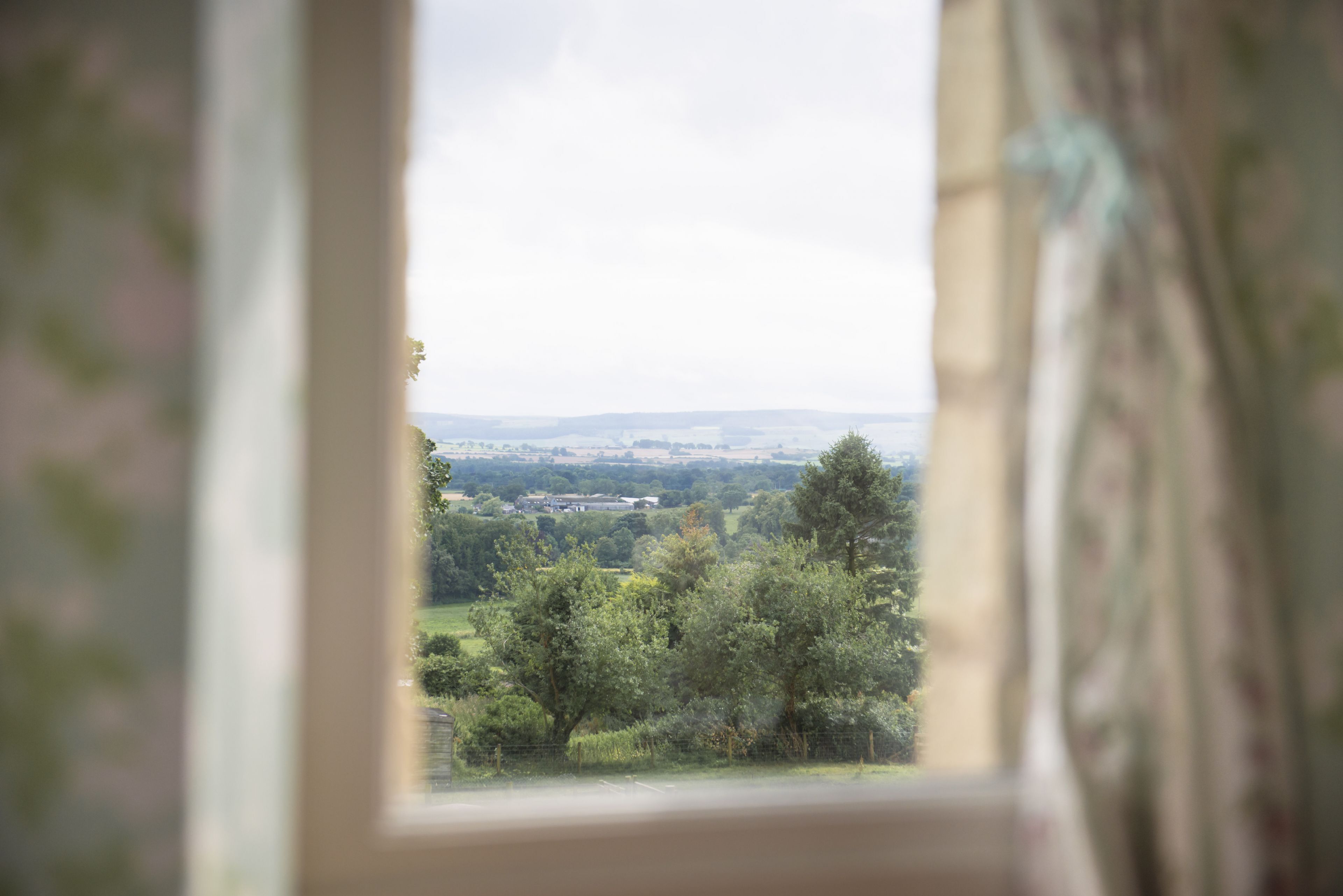 Une vue à travers une fenêtre, avec un rideau, qui révèle un paysage composé d’arbres et de bâtiments éloignés.