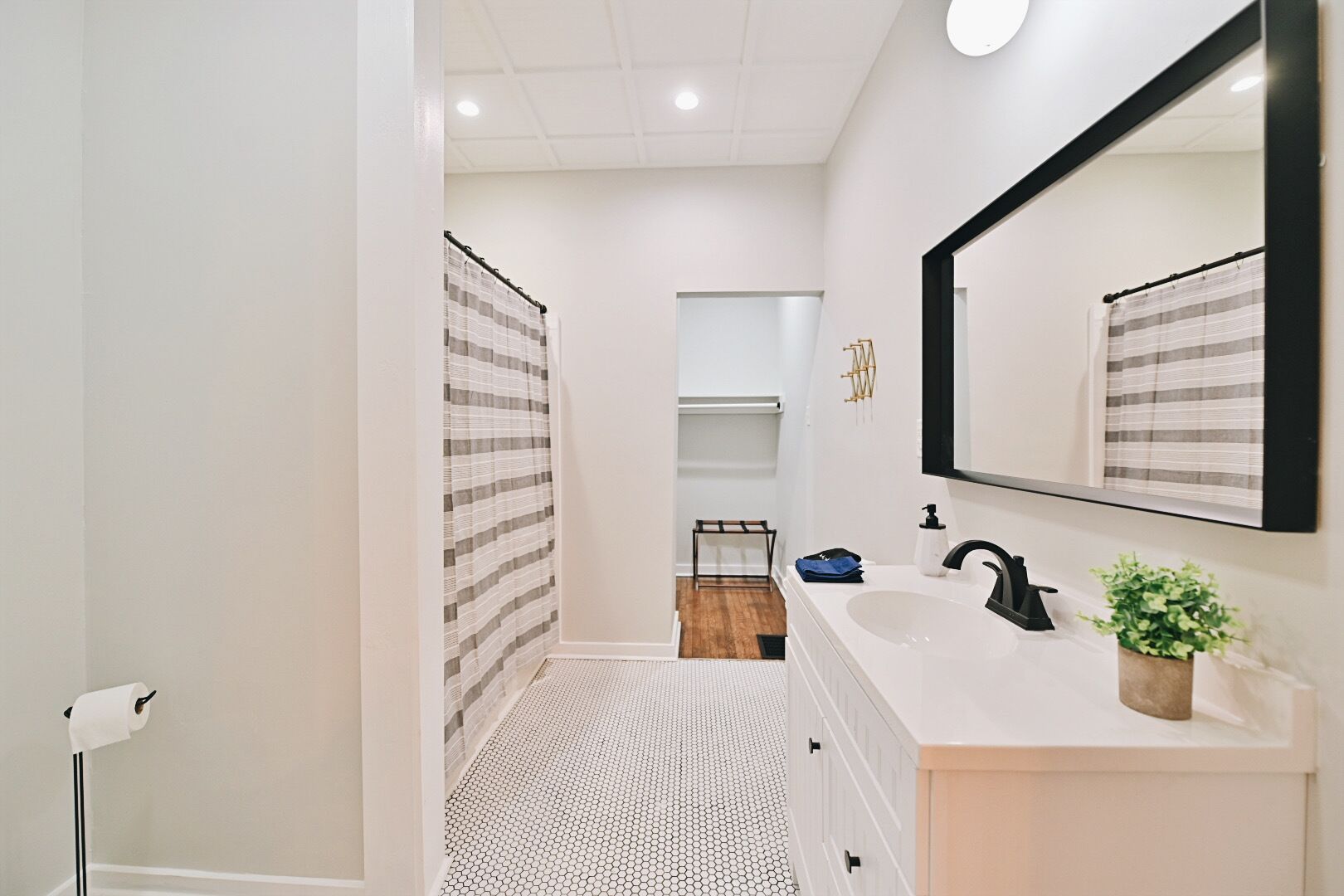 A modern bathroom with a white sink, black faucet, and a large mirror. There is a striped shower curtain and a small plant on the vanity.
