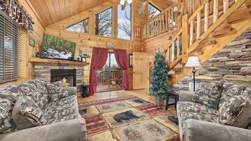 L’intérieur d’une cabane en rondins, avec une cheminée, deux canapés, un sapin de Noël et un escalier.
