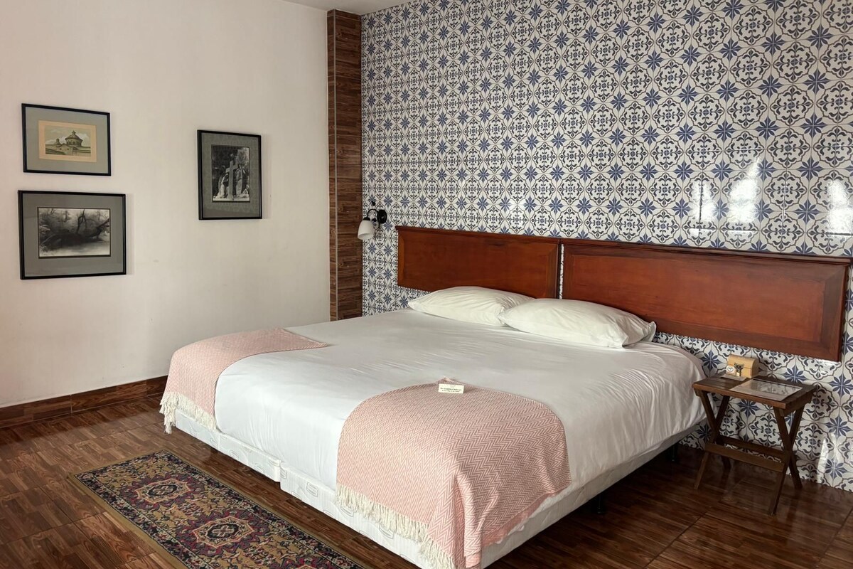 Une chambre à coucher avec un grand lit, une table de chevet, un papier peint à motifs et des tableaux encadrés accrochés au mur.