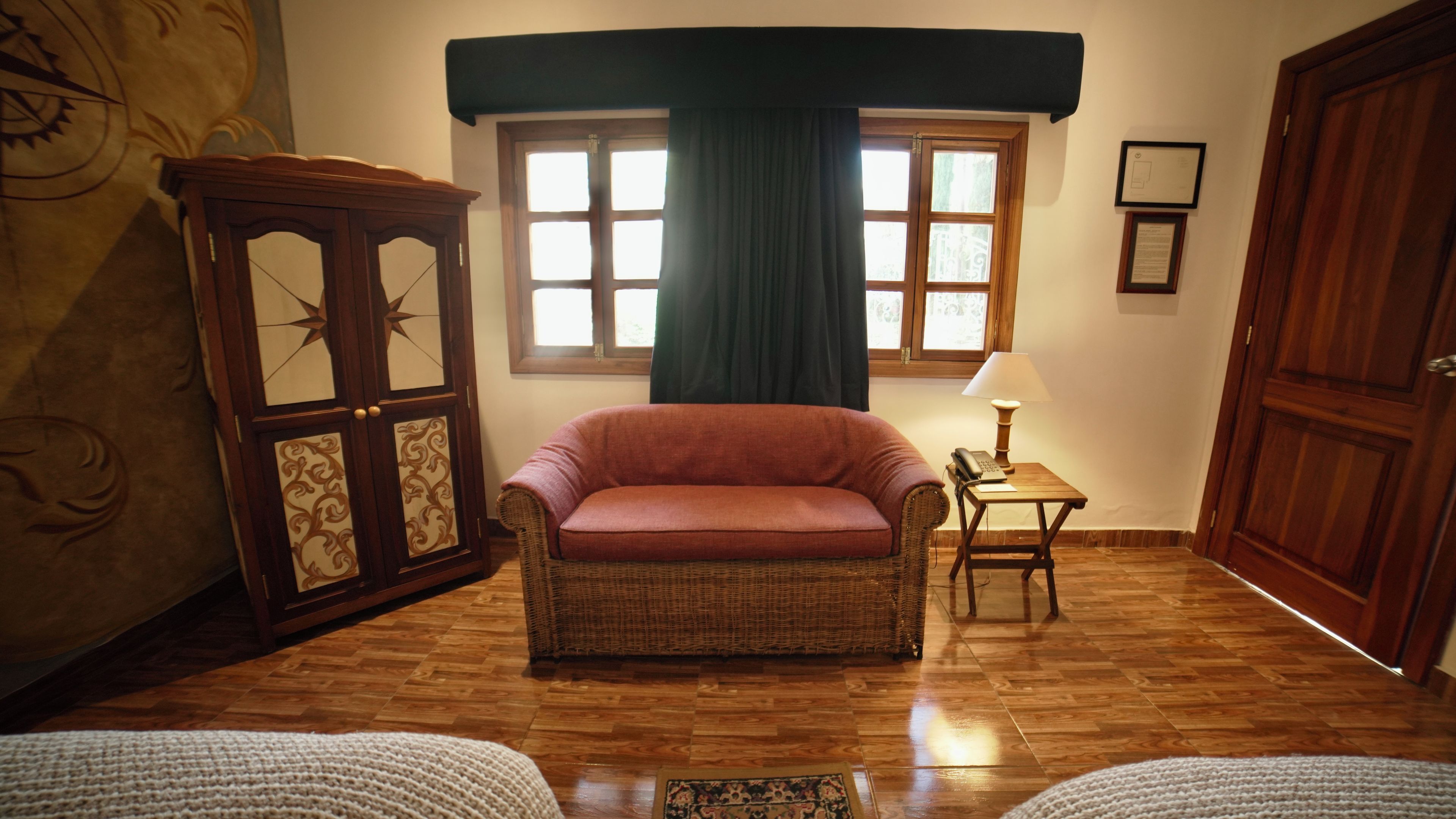 A room with a wicker armchair, a small table with a lamp, a wooden cabinet, and a large window with curtains.