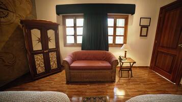 A room with a wicker armchair, a small table with a lamp, a wooden cabinet, and a large window with curtains.