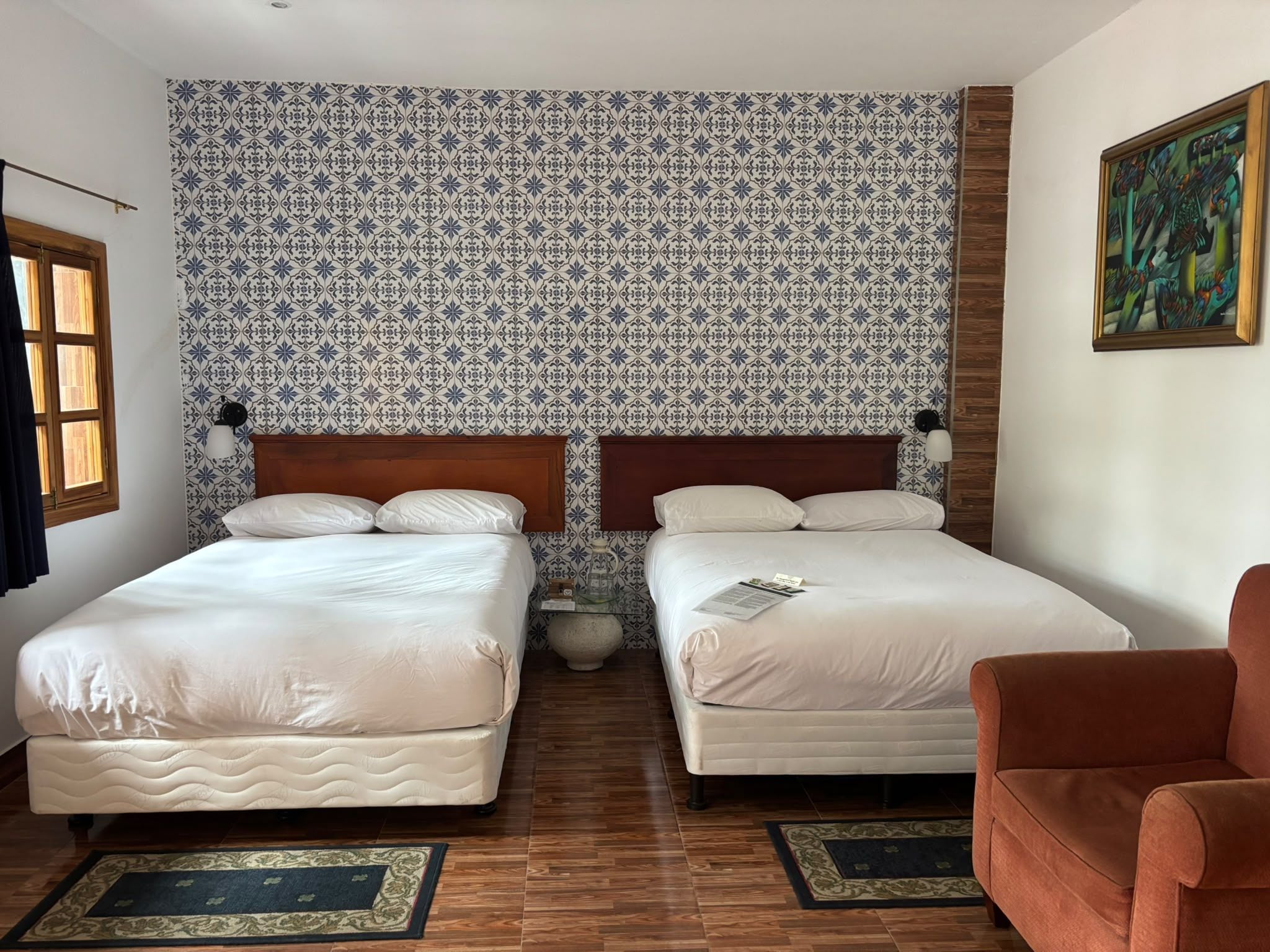 Hotellihuone, jossa on kaksi sänkyä, puulattia, kuvioitu seinä, taulu ja ruskea nojatuoli.