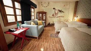 Habitación de hotel con piso de madera, un escritorio rojo con teléfono, sofá con cojines, cama con manta blanca y pared con un reloj y un mural de Charles Lindbergh.