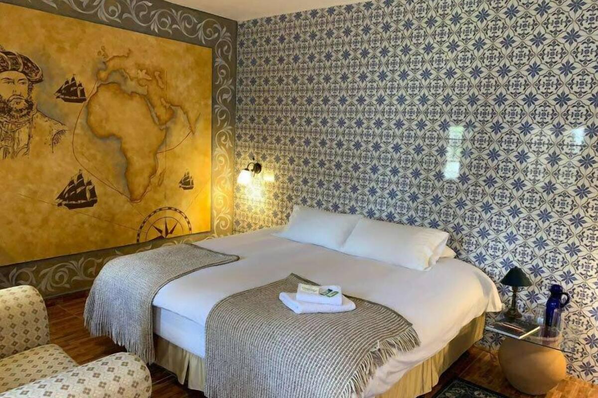 A hotel room with a large bed, a map on the wall, patterned wallpaper, and a small table with a lamp.