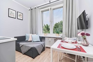 En moderne stue med en sofa, et spisebord dækket til to personer, et fjernsyn ophængt på væggen og et vindue med gardiner.