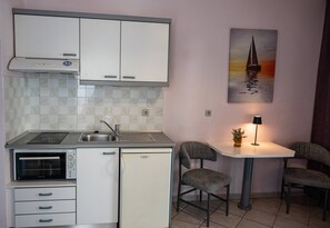Une cuisine compacte équipée d’un four encastré, d’un réfrigérateur et d’un évier. On y trouve une petite table à manger avec deux chaises et une lampe. Un tableau représentant un voilier est accroché au mur.