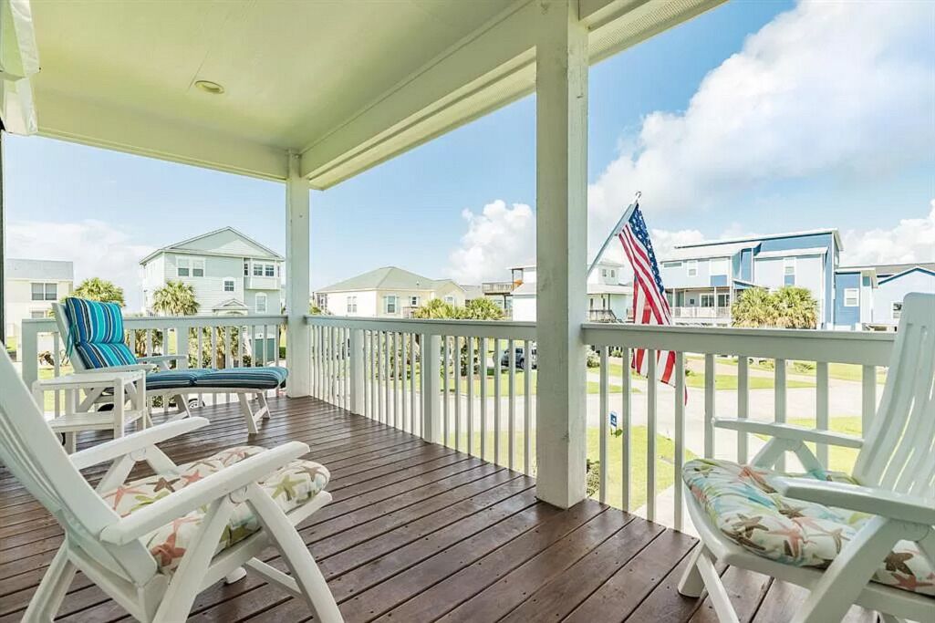 A covered porch with white railings and wooden flooring, featuring several white lounge chairs with floral cushions.
