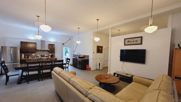 Ein modernes Wohnzimmer mit einem beigefarbenen Sofa, einem Essbereich mit Holztisch und -stühlen, einer Küche mit weißen Schränken und einem an der Wand befestigten Fernseher.