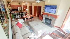 Una sala de estar con sofá, reposapiés y chimenea. Hay un comedor con mesa y sillas. Un televisor está montado en la pared.
