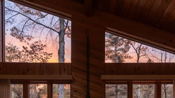 Una habitación acogedora con chimenea, dos sillones y grandes ventanales con vistas a un bosque.