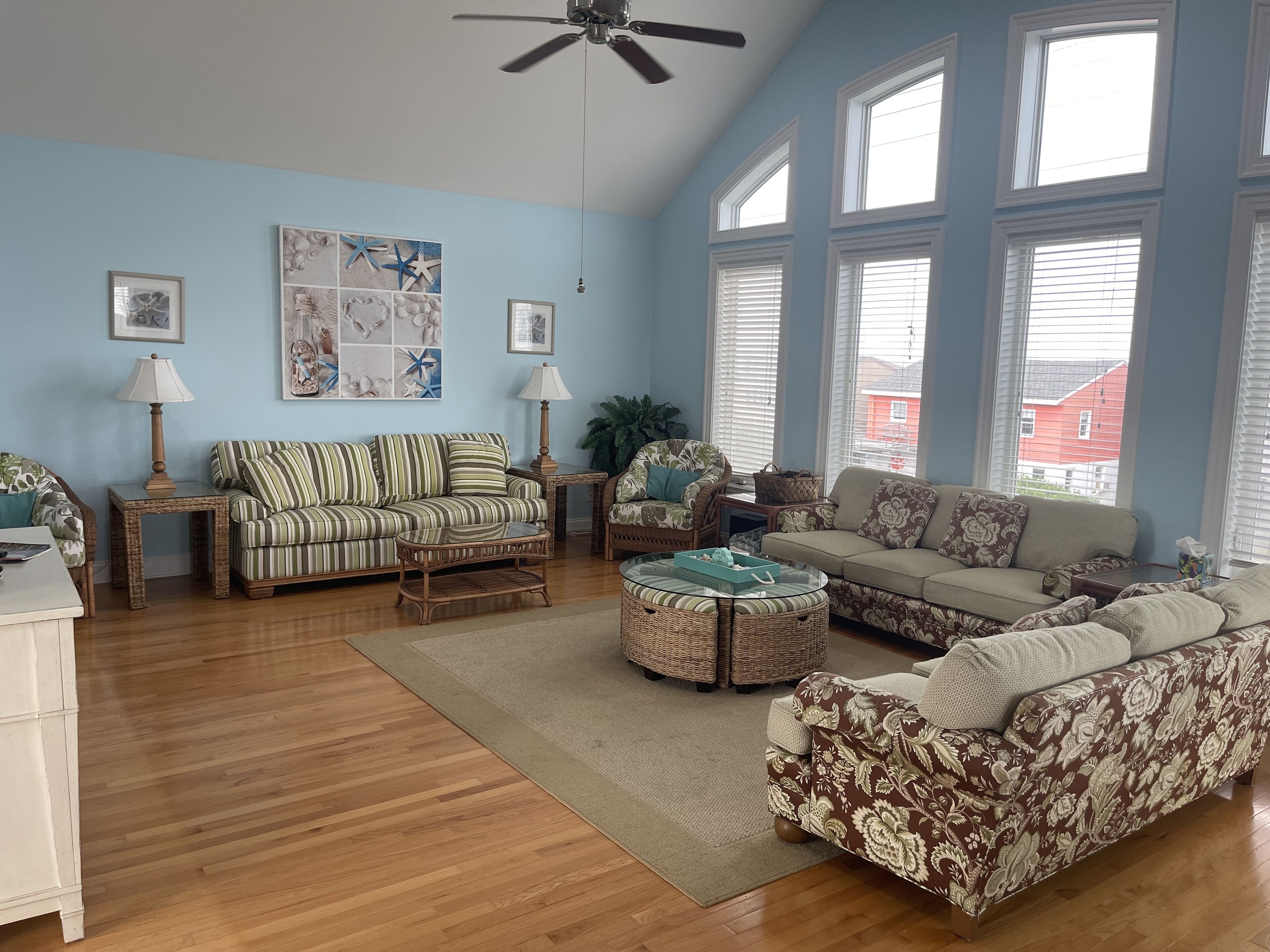 A living room with a high ceiling, large windows, and a variety of seating options including a striped sofa, a floral-patterned sofa, and a round wicker coffee table.