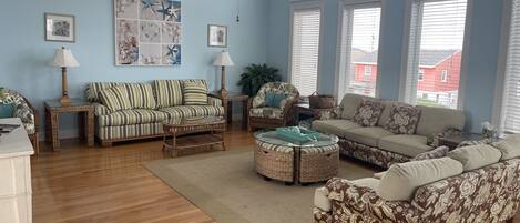 A living room with a high ceiling, large windows, and a variety of seating options including a striped sofa, a floral-patterned sofa, and a round wicker coffee table.