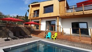 A modern two-story building with a pool, outdoor seating, and a wooden deck. - Villa Le Repère, quiet area, air-conditioned with swimming pool (SAINT AYGULF)