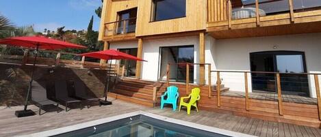 A modern two-story building with a pool, outdoor seating, and a wooden deck.
