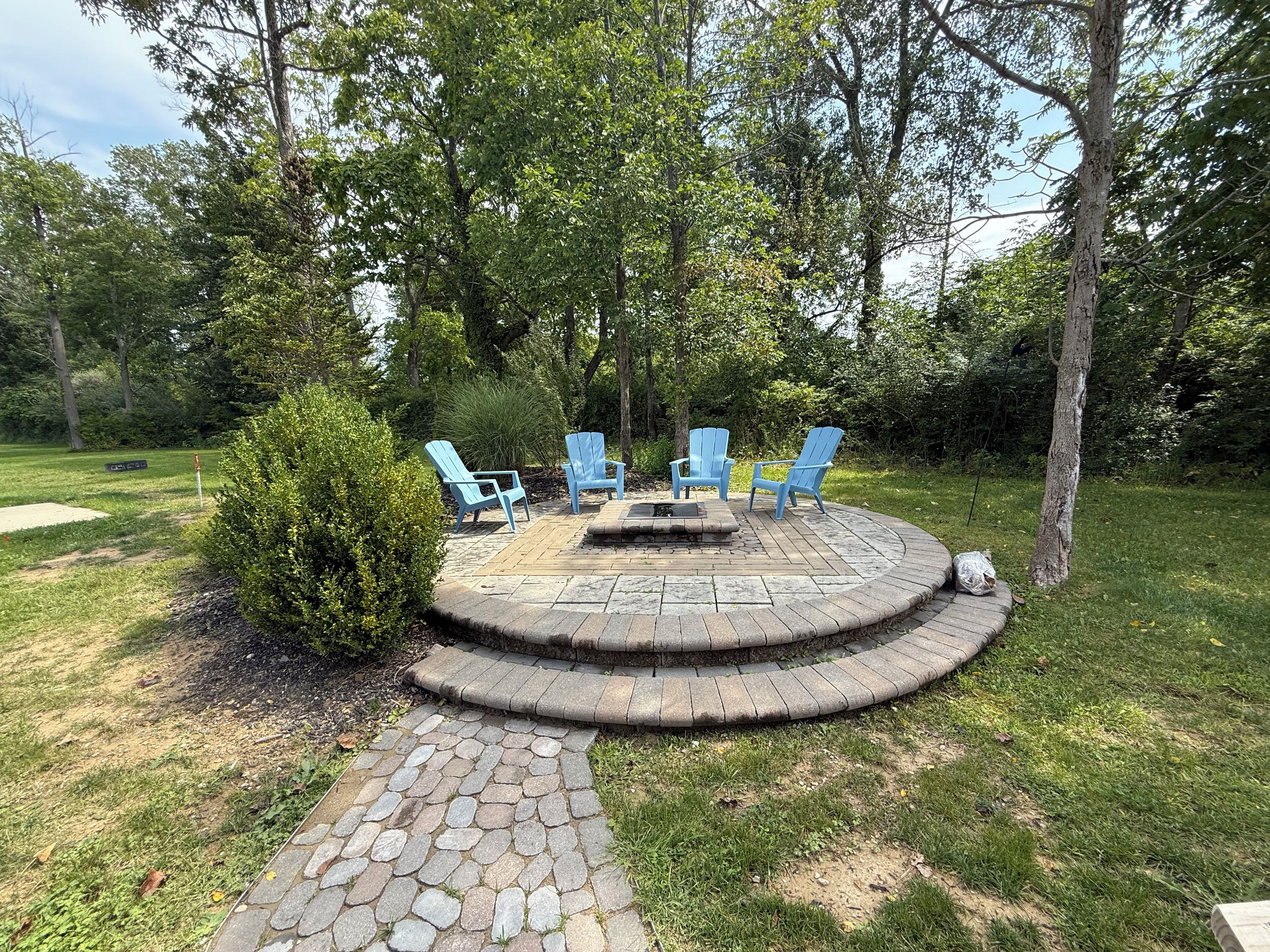 Un espace extérieur circulaire aménagé avec des chaises bleues et un foyer, entouré d’un chemin de pierre et de verdure.