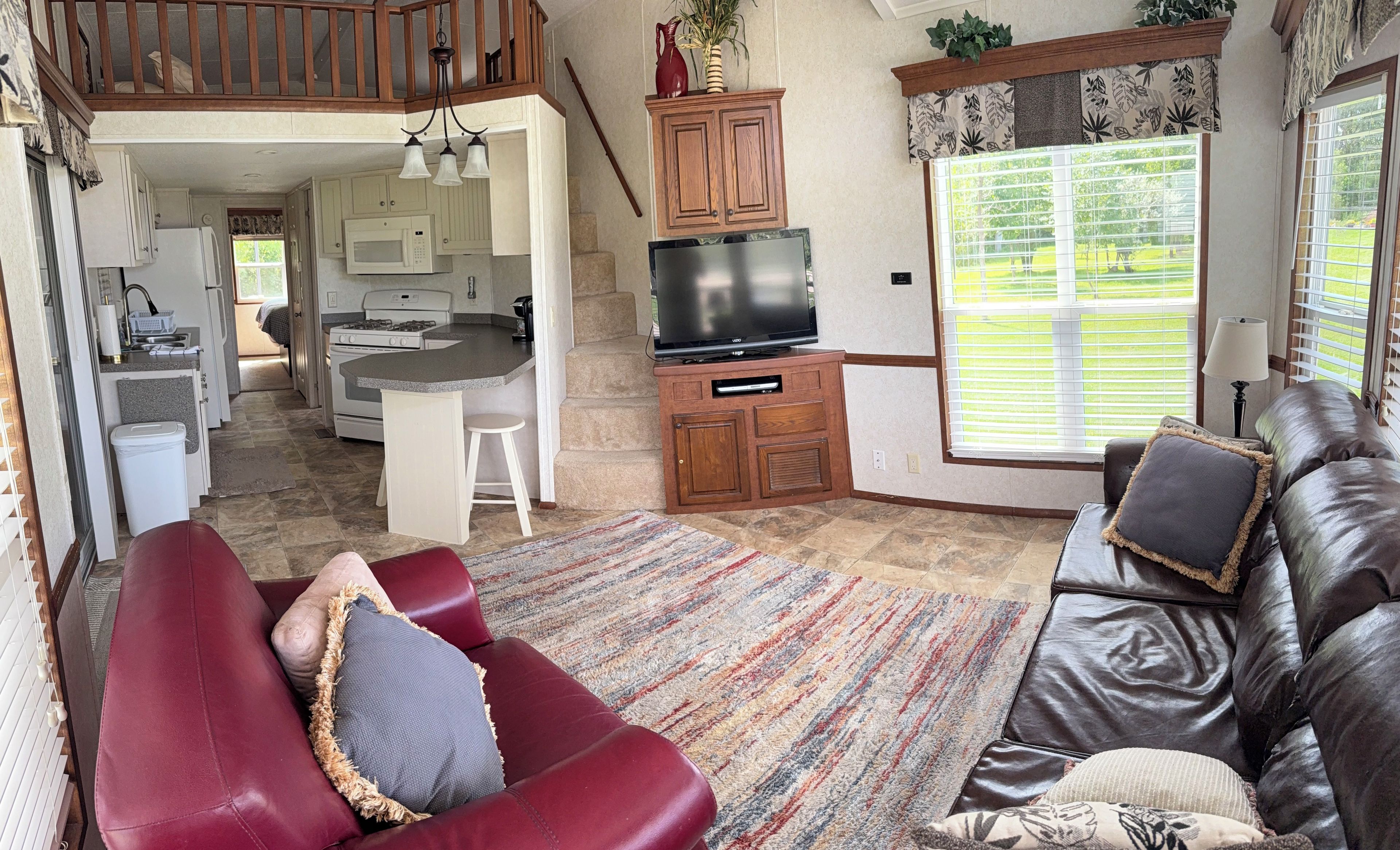 A living room with a leather sofa, a red couch, and a television. There is a kitchen area with a microwave and a stove.