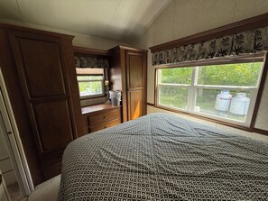 Un dormitorio con una cama grande, un armario de madera, un ventanal con vistas a follaje y una ventana con contenedores de almacenamiento.