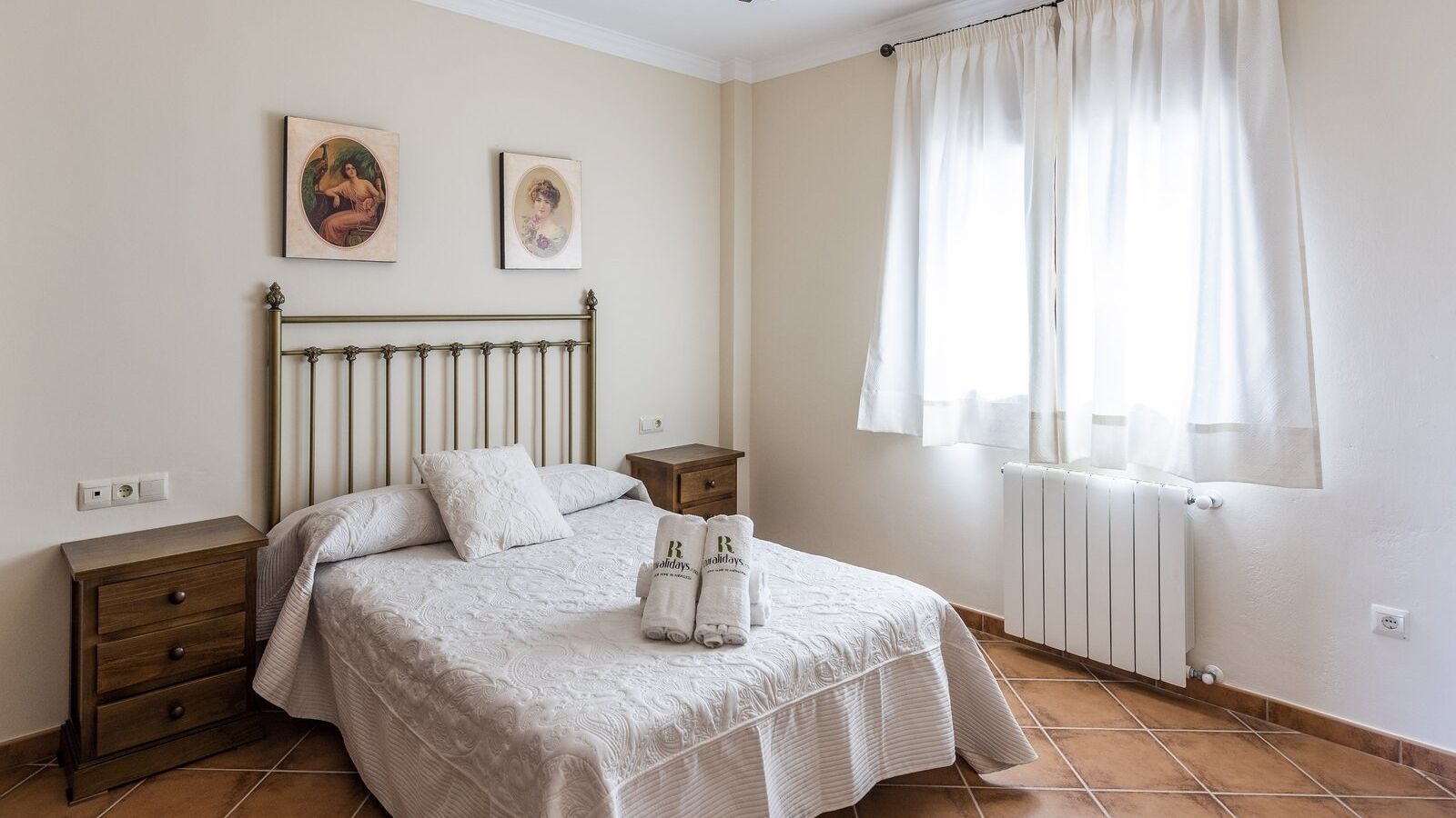 A bedroom with a bed, two nightstands, a radiator, and two framed pictures on the wall.
