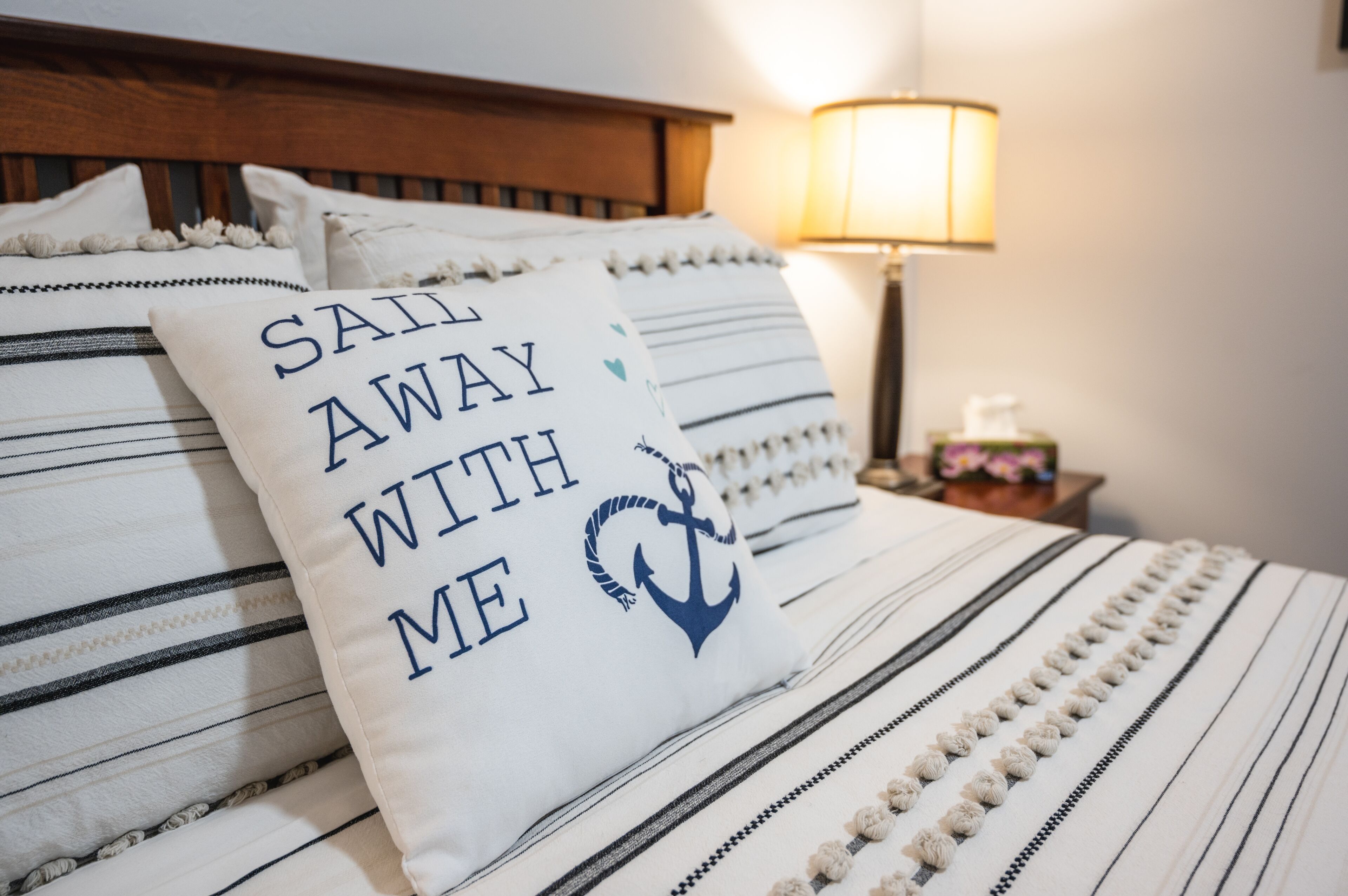 Een bed met gestreepte lakens en een kussen met de tekst 'Sail Away With Me' en een ankersymbool.