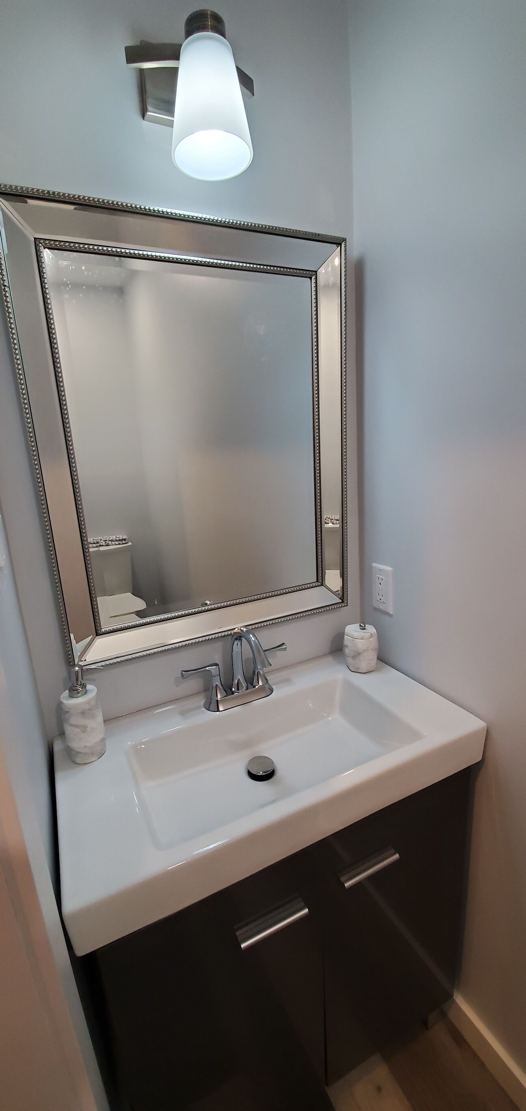 A bathroom with a white sink, dark cabinets, a large mirror, and a wall-mounted light fixture.