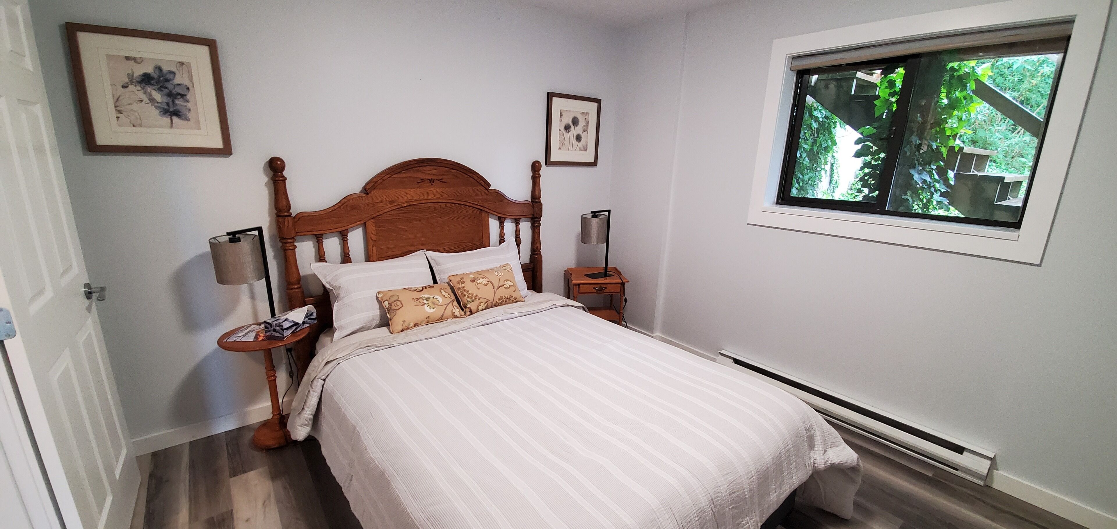 A bedroom with a wooden bed, white bedding, a bedside table with a lamp, and a window with a view of greenery.